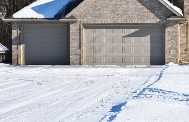 Snow-covered concrete driveway leading to a two-car garage at a residential home during winter, showing tire tracks and freeze-thaw conditions common in Illinois.