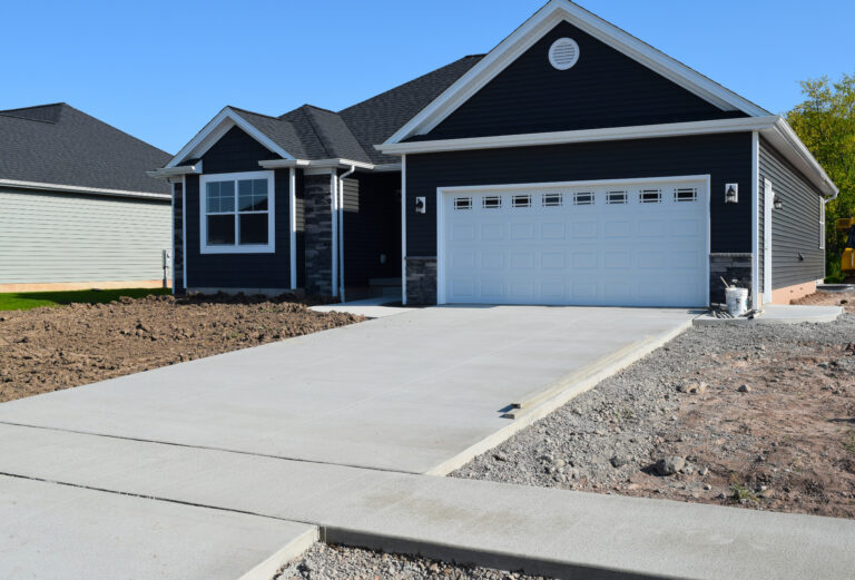 Modern residential concrete driveway leading to a dark blue suburban home with a white garage door, freshly poured smooth concrete finish, new construction landscaping, and clean curb appeal on a sunny day.
